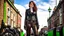 Placeholder: full-height portrait of a woman with straight shoulder-length auburn hair, with metal arms and legs, dressed in leather trousers, and a waistcoat, in a Victorian street next to a steampunk motorbike blue sky