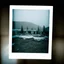 Placeholder: A polaroid photo of an odd room with structures. A transparent tarpaulin covers an image resembling a ruined building. There is a group of fir trees in the background and a high hill in the distance. The surface below is in a pastel color. Fog is present.