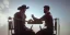 Placeholder: two cowboy men speaking while seated on a table in a room, bright light, high exposure