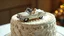 Placeholder: A hyper-realistic, detailed photo of a massive, elegant single-tier wedding cake. On top of the cake, there is a luxurious vintage open-top convertible toy car, made of fondant or edible sugar. Inside the car, sitting happily, are a tiny bride and groom doll. The cake is beautifully decorated with intricate white and silver icing, sugar pearls, and delicate floral patterns. The scene is lit with soft, warm studio lighting, creating a magical and joyful atmosphere. Shallow depth of field, focusin