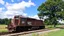 Placeholder: a brown train engine with the number 20 on it. The engine is a diesel locomotive with the word "NCCO" written on the side in white letters. It is sitting on a railway track with a tree on the right side and a grassy field on the left side. The sky is blue with white clouds and there are trees in the background. The train appears to be in motion, as if it is moving along the track. The overall mood is peaceful and serene.