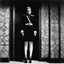 Placeholder: [Le Cabinet de Curiosités by helmut newton] The creature's immense almond-shaped eyes glow with an inner light, revealing a depth of ancient wisdom and unfathomable mystery. As it moves through the chamber, Clad in a Japanese-inspired ensemble of red, black, and white, the being wears a high kilt and thigh-high white stockings, adding a touch of ethereal allure to its reptilian form. A skeletal rib cage and massive vertebrae protrude from beneath the cybernetic exoskeleton, hinting at a fusion o
