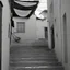 Placeholder: Calle de un pueblo de una isla italiana en verano, con escalinata, fotografía real, fotografía realizada con cámara Leica y objetivo de 50 mm, siguiendo estilo de la serie 'Ripley' emitida en Netflix, fotografía en blanco y negro, virada tonos años 50