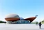 Placeholder: 3D representation of architectural wonder, with a concrete design and matte reddish glass that contrasts with the light blue sky, emphasizing organic movement. Its design represents an ant with a bulbous tail and membrane wings with solar panels, its tail is made of concrete and glass. It stands out from pedestrians, creating a sense of scale. In open space, its use is a public place and as a great viewpoint in the mountains Combination of practicality and artistic expression in architecture