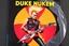 Placeholder: Record sleeve for 80's metal band text "DUKE NUKEM" for album text "GREATEST BLASTS", Duke Nukem blonde high buzz cut character dramatically jamming on an electric guitar that is styled like a ICMB missile, background is a radiation symbol, over the top Duke Nukem aesthetic, digital photograph, vinyl record partially pulled from sleeve