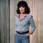 Placeholder: Scene from a color 1968's British secret agent TV show, a 165 pound middle aged woman who looks like Sherri Martel is wearing jeans and boots, she has on very heavy eye makeup and her mouth is open. She is standing up with her arms at her sides, her knees are buckled and her eyes are crossed as they close and roll. She looks very defeated and humiliated. She is standing in a colorful living room.