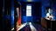 Placeholder: Image is a painting titled 'Blue Room' from 1960, measuring 28 by 50 inches. It depicts an interior scene with a predominantly blue color palette. The room features a patterned blue wallpaper with red floral designs. On the left, an open door reveals a bright light source, casting a warm glow into the room. The floor is a reflective blue, enhancing the room's cool tone. A large, oval mirror hangs above a simple fireplace on the right wall, which is adorned with a subtle geometric pattern. To the