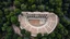 Placeholder: an aerial view of an ancient amphitheater. The structure is semi-circular with stepped seating that curves around a central stage area. The amphitheater seems to be in a state of partial ruin, with some sections clearly showing their age. The surrounding area is densely populated with trees, giving a sense of isolation and natural beauty. The overall scene reflects a blend of historical architecture and natural surroundings.