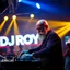 Placeholder: A dj on a night rave club standing mixing songs , he looks like walter white kinda, bald with beard. strobes and blue yellow light, a big sign with "DJ ROY" in the background, this is a album cover for his dj roy essentials album.