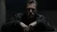 Placeholder: In the photo, we see a man sitting in the dark, looking at the camera with a cold gaze. The man is dressed in black and seems uninterested in what is going on around him.