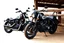 Placeholder: a motorcycle(harley davidson pan america 1250) next to a different motorcycle(Indian Springfield Dark Horse) , next to a horse who's tied by the reins to a wood railing. white background. dirt ground. silkscreen style image