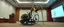 Placeholder: pinhole cam, airbrush 3d rendering, on tv screen:A five-second cinematic single take in a clean industrial presentation hall. A bizarre mechanically plausible tracked bicycle machine stands beside a presentation podium, designed to look like a giant mechanical praying mantis. Two large bicycle wheels form its main body and four thin articulated mechanical legs support it. Two long folded mechanical front arms resemble mantis forelegs, huge head and arms