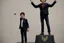 Placeholder: The boy is standing on a stylized podium, raising his right hand victoriously in the air, with his fingers forming a "V" sign. His left hand holds a symbolic representation of his achievement, which could be a miniature golden cup, a shining star, or any object that reflects the context of the competition or event.
