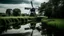 Placeholder: A black windmill with four sails reflected in a calm river, surrounded by a grassy field and a row of trees under an overcast sky