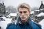 Placeholder: a Russian young man with handsome features and blonde hair, he is wearing a blue millitary uniform, he is standing in the snow on gray rocks, it is winter and snowflakes are falling, there is a destroyed village in the background