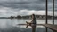 Placeholder: Woman sitting on a jetty with her feet in the water of the lake, in the background you can see a house of modern architecture that is reflected in the lake, the sky threatens storm