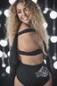 Placeholder: extremely tiny, thin, beautiful 21-year-old Sheena Greane with long, wavy curly blonde hair, wearing black strapless sports bra and cotton shorts, smiling a happy smile, extremely giant, oversized, humongous orbs, pitch black background, professional quality studio 35mm 8x10 uhd photograph,