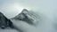 Placeholder: a massive sheer snowy mountain cliff with very sparse vegetation scaling vertically into the sky, partially obscured by dense clouds and mist. the borders — top, bottom, left, and right — fade smoothly into thick fog, while the center reveals the steep, rocky cliff face with fine texture and detail. atmospheric lighting, cinematic composition, natural colors, high contrast between fog and stone.
