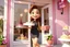 Placeholder: sign "Mon Petit" 3D video game character elegant young woman enthusiastically and cheerfully comes out of a dessert store, cake box in her hand, in the window cakes, cookies, macarons and flowers in sunshine