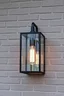 Placeholder: An industrial-style outdoor wall light fixture mounted on a gray brick wall. The light has a rectangular black metal frame with clear glass panels, enclosing a tall filament bulb that emits a warm glow. The texture of the brick wall enhances the modern, minimalist design of the fixture, creating a cozy and inviting ambiance