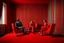 Placeholder: Un hombre vestido con traje, un hombre con pijama rojo, y una mujer vestida con traje, sentados en sillones, en una habitación de suelo de baldosas con líneas zigzagueantes, la habitación la recorre una gran cortina roja, fotografía realizada con cámara Leica y objetivo de 50 mm. Estilo serie de televisión Twin Peaks