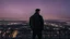 Placeholder: An Englishman in a bomber jacket standing to one side of a tall building looking across a city after sunset