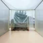 Placeholder: A Hasselblad H6D-400c photo of an odd room with structures. A transparent tarpaulin covers an image resembling a ruined building. There is a group of fir trees in the background and a high hill in the distance. The surface below is in a pastel color. Fog is present.