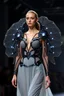 Placeholder: Donna in passerella con Struttura scultorea semi-rigida, tipo esoscheletro che si apre e si chiude Materiali: stampa 3D bio-based, tessuti fotocromatici, rete metallica ultra sottile Dettagli: Reazioni ottiche alla luce, conchiglia futuristica che pulsa col corpo