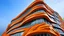 Placeholder: A building featuring a design of steel ribbons on an orange facade, set against a gray sky. The ribbons are arranged in waves, creating a dynamic and fluid appearance. The orange facade can be seen between the ribbons, creating a chromatic contrast that accentuates the building's unique design. The view is taken from a low angle, which emphasizes the height and scale of the building.