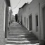 Placeholder: Calle de un pueblo de una isla italiana en verano, con escalinata, y arcos, tono decadente, fotografía real, fotografía realizada con cámara Leica y objetivo de 50 mm, siguiendo estilo de la serie 'Ripley', fotografía en blanco y negro, virada tonos años 50