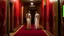 Placeholder: Two people are depicted in a hallway. Both are wearing loose, light-coloured, almost white dresses, with slightly tattered edges, in a loose, romantic style. Their expressions are melancholic, wistful or pensive. They are walking down a red-carpeted hallway; the effect of the carpet is soft, deep red. The hallway is lined with light brownish-green walls and doors. A large, wilted sunflower rests on the stairs, its head down, its petals a vibrant yellow, contrasting with the dark-hued, almost bla