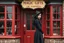 Placeholder: Photorealistic image of a slender, young woman with dark hair, in a witchy, black dress, no hat, leaning back against a shop door with red-painted, large, multi-paned windows on either side of a red-painted multi-paned door. Walls made of brick. The shop is selling magic gifts and trinkets. Large sign over the door.