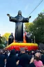 Placeholder: 200 ft tall floating cartoonish jesus parade balloon pulled by multiple grim reapers, multi colored, bright, vivid, psychedelic, high detailed, high gloss, insanely detailed, matte, horrorcore, lining the street, unsettling contrast, monument to death, live action shot grim reaper parade, pulling a jesus parade float