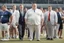 Placeholder: Overweight white politician wearing a suit; next to him a group of thin, muscular athletes in sports clothes; outside, sports field; realistic, photographic