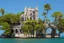 Placeholder: A futuristic ruined gothic building on an island floating over the sea with balconies, verandas, many arches, bridges, spires, paths, trees, dense foliage, spanish moss, ivy, blue sky, white clouds