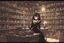 Placeholder: full-height shot of a young witch in a tight black short skirt, inside a large magic shop, sitting at a desk, shelving, bottles, windows