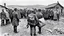 Placeholder: an man in a dark european, hungarian postman's uniform standing with his back to the camera with a large postman's bag on his left shoulder, he is only half visible from the european-hungarian villagers gathered around him, surrounded by men and women in vintage poor cloths. Some villagers are holding letters in their hands, sad and crying faces, a little boy staring at the postman, a crying little girl clinging to her mother's skirt, two women crying , high realistic, perfect photo