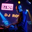 Placeholder: A dj on a night rave club standing mixing songs , he looks like the actor Bryan Cranston, bald man. strobes and blue yellow light, a big sign with "DJ ROY" in the background, this is a album cover for his dj roy analisentials album.