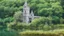 Placeholder: A ruined gothic stone building in a lake, balconies, verandas, arches, bridges, spires, stairs, trees, dense foliage, spanish moss, ivy, blue sky, white clouds