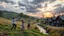 Placeholder: a group of young people are dancing to camera in village center squre over high grassy hills,a small fall and river and wild flowers at river sides, trees houses ,next to Ripe wheat ready for harvest farm,windmill ,some people standing looking the dancers,a few village local shops ,cloudy sun set sky