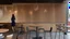 Placeholder: A modern cafe interior with a woman standing near a table. The walls and ceiling are covered in horizontal and vertical wooden slats, creating a warm and textured atmosphere. The floor is a polished gray tile. Several round tables with modern chairs are arranged in the space. Pendant lights hang from the ceiling, casting a soft glow. The overall style is minimalist and sophisticated, with a focus on natural materials and clean lines.