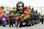 Placeholder: cartoonish jesus parade balloon pulled by multiple grim reapers, multi colored, bright, vivid, psychedelic, high detailed, high gloss, insanely detailed, matte, horrorcore, lining the street, unsettling contrast, monument to death, live action shot grim reaper parade, pulling a jesus parade float