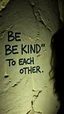 Placeholder: A hand writing On cave wall written “BE KIND “ “ TO EACH OTHER “ an old man with bear and king hair died by the cave wall , a dim lighting great quality picture with primitive style