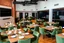 Placeholder: 4k Image depicts a modern, spacious dining area with a contemporary style. The layout features several rectangular tables arranged in rows, each set with green velvet chairs. The tables are elegantly set with plates, cutlery, and small floral centerpieces. The room has a warm ambiance, highlighted by wooden flooring and exposed brick walls. On one side, a large flat-screen television is mounted on a white wall panel. The opposite side showcases a sleek, black kitchen area with built-in appliance