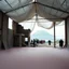 Placeholder: A Hasselblad H6D-400c photo of an odd room with structures. A transparent tarpaulin covers a ruined building. There is a group of people in the background and a high hill in the distance. The surface below is in a pastel color. Fog is present.