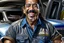 Placeholder: Fotorealistisch Danny Trejo breit grinsend mit offenem Haar in ölverschmiertem Mechaniker Overall fährt Muscle Car