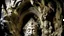 Placeholder: A large stone cave carved into a rock formation and abundantly carved in a baroque or rococo style, with a human face with an elaborate headdress or crown, surrounded by intricate decorative elements, human and organic figures, mythological animals, leaves and plants. The face appears to be that of an older man with a serious expression.