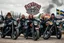 Placeholder: a very hard and tuff motorcycle young gang with Keltic hair , not old, posing with their Harley Davidsson bikes, some of them holding guns and shotguns, a logo sign with "FALKÖPING HD", "FEARLESS SINNERS" Skulls and bones, skeletons and fire, Swedish town Falköping in background, Swedish flags on bikes, Simon Stålenhag style