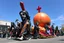Placeholder: cartoonish jesus parade balloon pulled by multiple grim reapers, multi colored, bright, vivid, psychedelic, high detailed, high gloss, insanely detailed, matte, horrorcore, lining the street, unsettling contrast, monument to death, live action shot grim reaper parade, pulling a jesus parade float