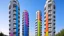 Placeholder: Tall cylindrical buildings dominate the scene, each constructed from light gray concrete. The structures are distinctive, punctuated diagonally by a series of circular windows, some partially open, contributing to the modern, industrial aesthetic of the image. Across the facade are vibrant, pixelated rainbow stripes, their colors shifting from red and orange at the bottom to blue and violet at the top. This striking optical effect adds a dynamic sense of movement and contrast to the otherwise mo